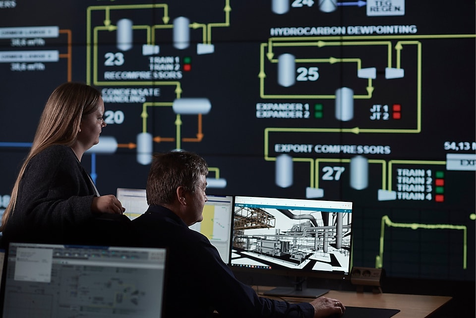 Two people in the office working on a computer, in front of a big screen showing production data.