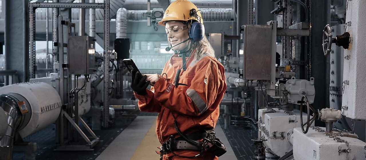 ung smilende kvinne kledt I verneutstyr jobber på en iphone ute på et anlegg.