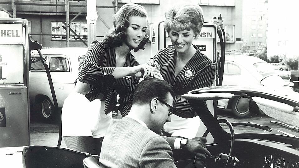 Historisk bilde i sort/hvitt: To kvinnelige Shell-ansatte gir service til en bilist-kunde i kjørebanen.