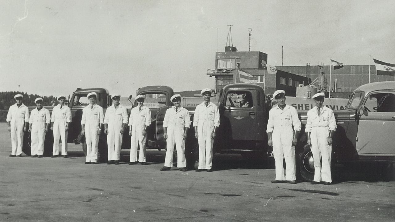 Historisk bilde i sort/hvitt: En gjeng med Shell-menn kledt i hvitt står foran en tankbil.