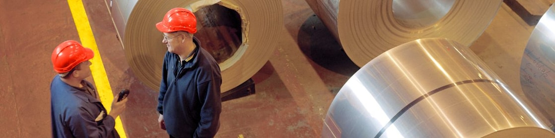 Two factory workers talking next to large rolls of steel